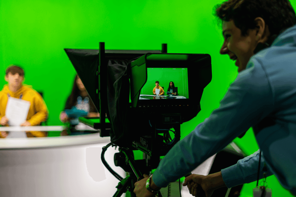 A person operates a camera filming two people sitting at a desk in front of a green screen.