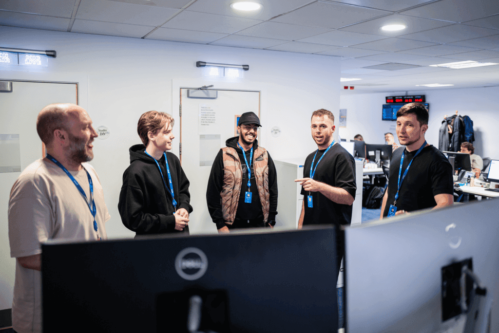 Five people stand in a circle in an office, wearing lanyards, and engaging in conversation. Computer monitors and office furniture are visible in the background.