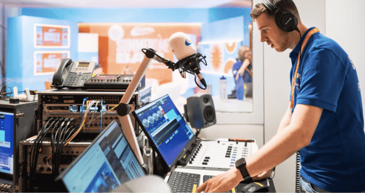 A man wearing headphones operates controls in a radio broadcasting studio, surrounded by equipment and screens.