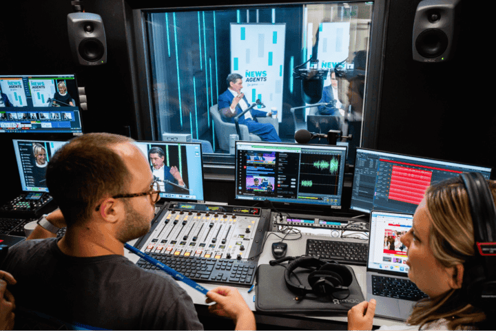 Two people operate audio equipment in a studio, while a man is speaking in a glass-enclosed booth labeled News Agents in the background.