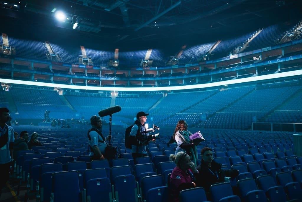 A film crew with cameras and equipment stands in the seating section of an empty, dimly-lit arena.