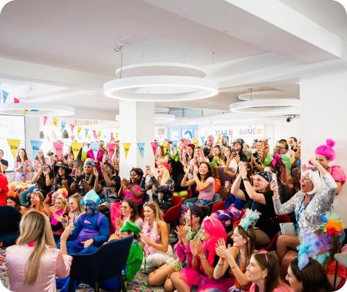 A large group of people in colorful, festive outfits sit and stand in a decorated room, clapping and cheering.