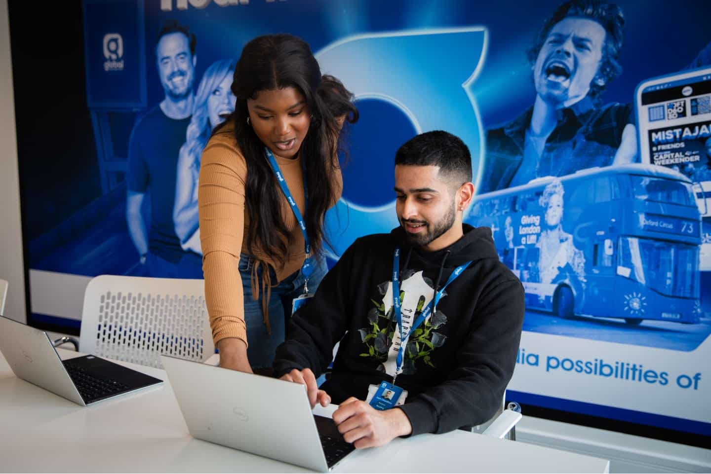 Two colleagues in an office setting collaborate on laptop work, with a promotional backdrop featuring a bus and public figures. Both wear identification lanyards.