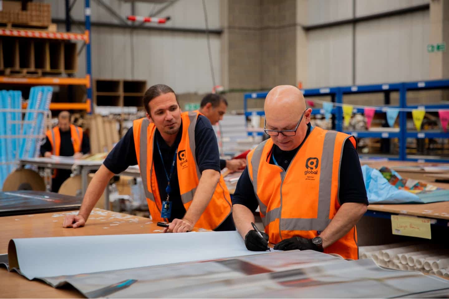 Two workers in orange vests handle large sheets of paper at a busy warehouse, surrounded by industrial supplies and shelving.