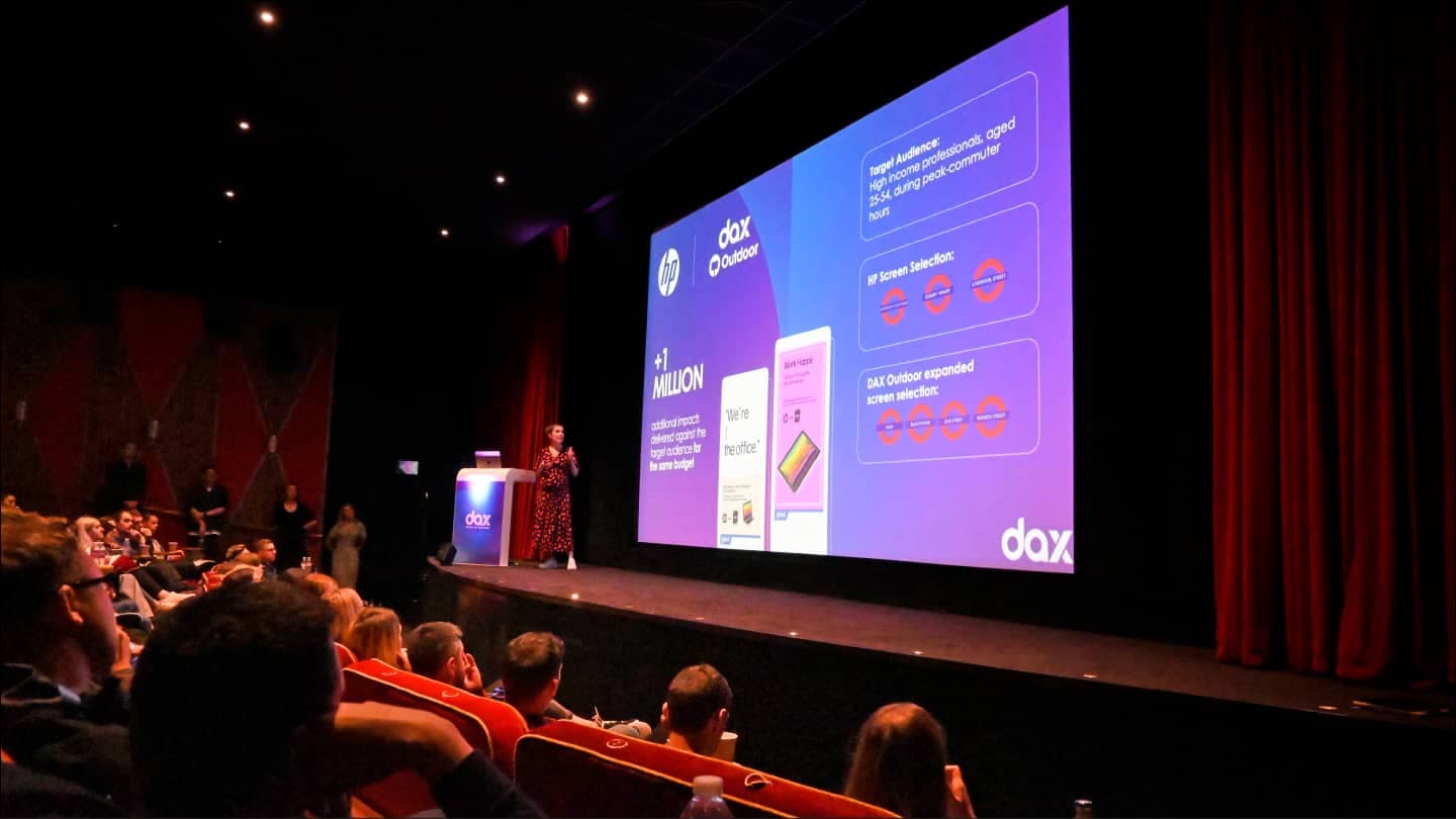 A person presents on stage in a theater, displaying a large slide with digital advertising statistics to an audience.