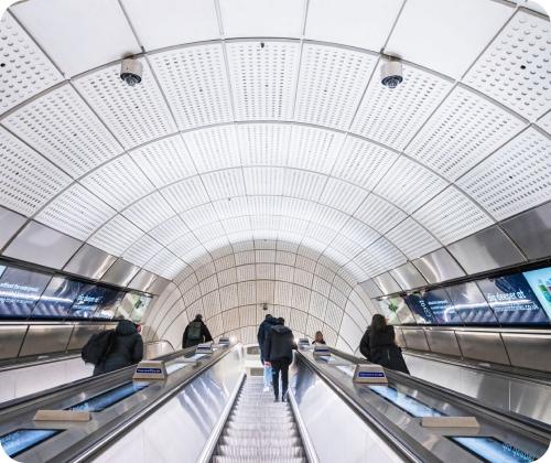 People stand on escalators in a modern underground station with a curved, tiled ceiling.