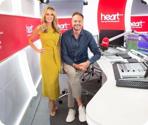 Two people in a radio studio, one sitting and one standing, with microphones and sound equipment around them.