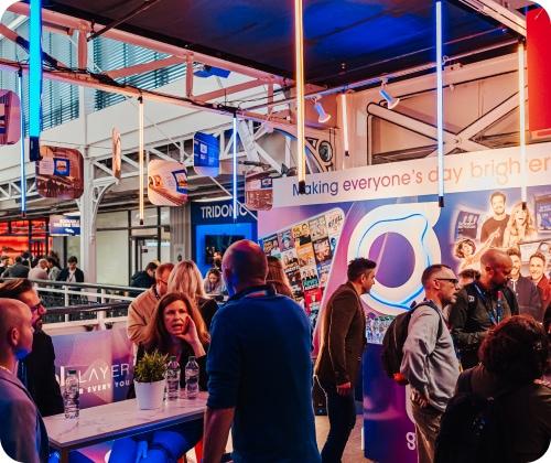 A group of people interact in a brightly lit booth at an indoor event. The booth features colorful signage and a display of various materials.