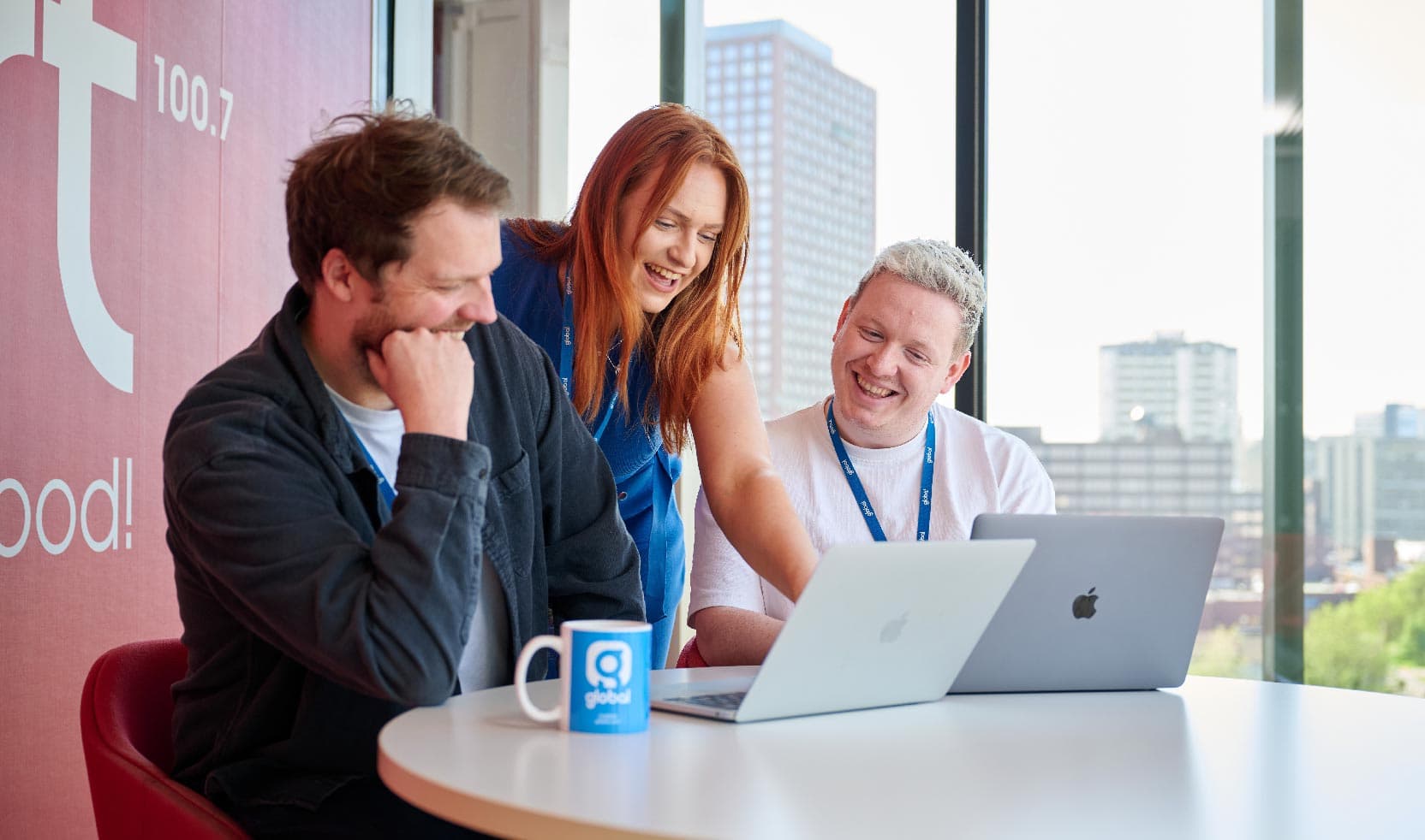 2 men sat at a table in a meeting room casually looking at 2 laptop screens. 1 woman wearing blue is smiliing and leaning over whilst pointing at the closer laptop screen. They are in a small red meeting room that has a large window that you can see a skyline through.